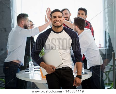 Picture of handsome businessman posing with his hand in pocket in office interior. Many business colleages or office workers showing victory of teamwork on background.