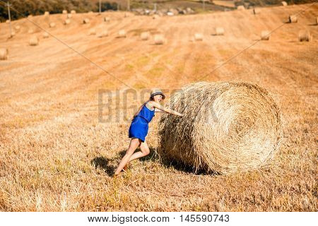 Young woman in blue dress pushing hay roll on the hayfield in Tuscany in Italy