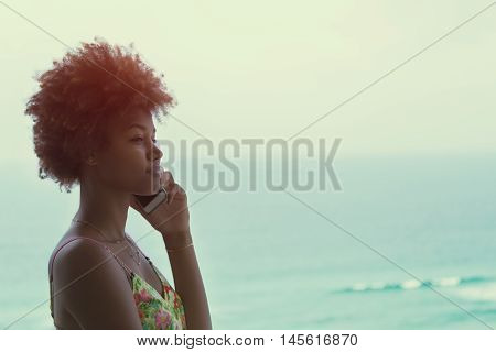 Young black pretty attractive female with curly hair speaking on a mobile phone, standing near tropical ocean with small waves, sunny bright summer day, silhouette, vintage color filter