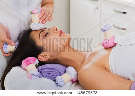 Herbal ball massage in ayurveda spa. Female massagist with young woman patient in wellness center. Professional cosmetologist make health therapy to beautiful indian girl in beauty parlor.