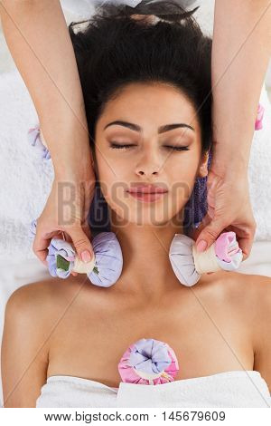 Herbal ball massage in ayurveda spa. Female massagist with young woman in wellness center. Healthcare therapy to beautiful indian girl in beauty parlor, top view of face with eyes closed