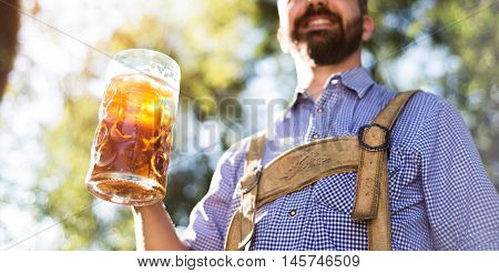 Unrecognizable young hipster man in traditional bavarian clothes holding a mug of beer. Oktoberfest. Sunny summer garden.