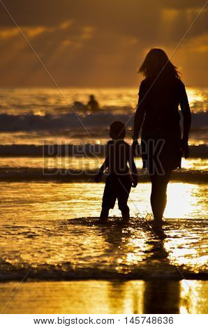 Mother and son walking on the beach in the surf at sunset silhouette.