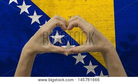 With a stylized Bosnian flag background an anonymous person's hands being held in the form of a heart, symbolizing love and patriotism for Bosnia and Herzegovina.