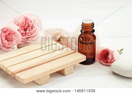 Rose essential oil bottle, fresh pink flowers, wooden surface for product display. Holistic beauty care. Creamy pastel tones, soft focus.