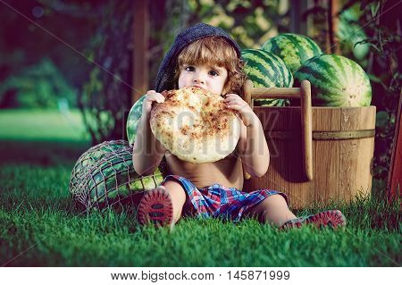 Little boy sitting on the grass near the pile of watermelons bite the big cake. Closeup portrait in retro style