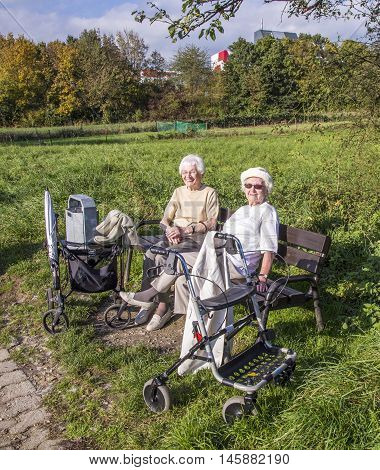 Two Elderly Ladies Enjoy The Sun At A Bench And Got There With A Rollato