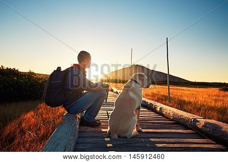 Man with dog on the trip in the mountains. Young tourist and his dog watching the amazing sunrise. Footbridge across the moorland and Snezka - the highest mountain in the Czech Republic.