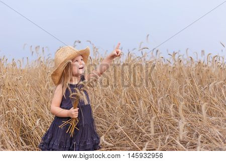 Baby girl in a big straw hat standing in a wheat field with a bunch of ears and hand points in the sky birds.