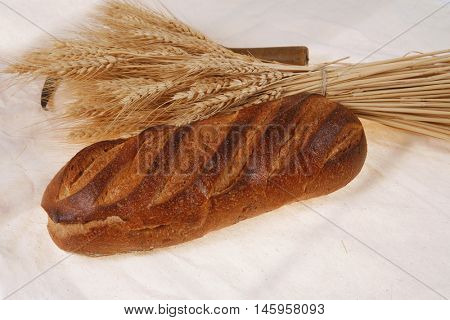 bunch of mown wheat ears with vintage handmade reaper hook sickle and rye dark white french fresh bread loaf on light linen tablecloth