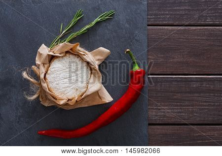 Cheese delikatessen closeup with chili pepper on black stone surface and wood. Camembert or brie circle in brown kraft paper decorated with basil, top view image