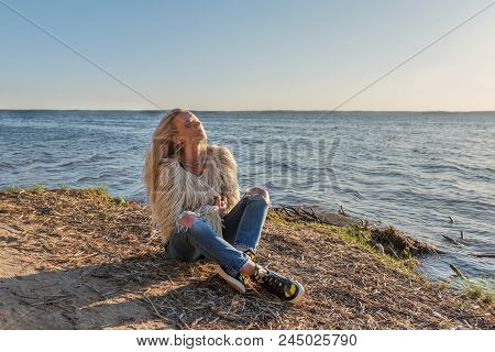 A Young Girl Sits Lounging On The Shore Of A Pond And The Wind Blows Her Blond Hair