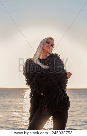 Blond Model In A Black Hairy Jacket Is In A Fashion Pose With Water And A Setting Sun On The Backgro