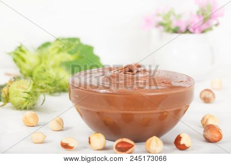 Homemade Hazelnut Spread In Glass Bowl. Hazelnut Nougat Cream. Violet Flowers In Small Vase In Backg