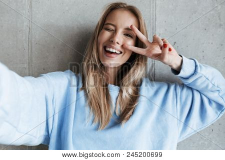 Picture of young cheerful happy pretty woman standing over grey wall background make selfie by camera showing peace gesture.