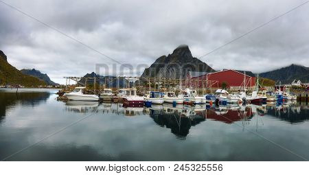 Fishing Boats Anchored At The Port Of Reine Village With Mount Olstind In The Background On Lofoten 