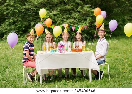 holidays, childhood and celebration concept - happy kids sitting at table on birthday party at summer garden