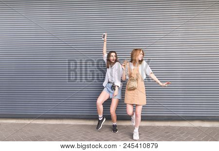 Two Fashionable Young Girlfriends Dance On A Gray Background. Portrait Of A Full-grown Two Girls In 