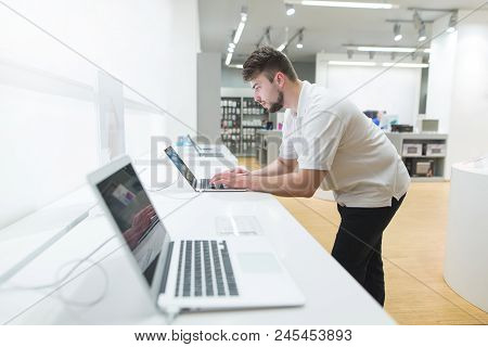 Man Uses A Laptop In The Computer Department Of The Technology Store. Showcase With Laptops. The Buy