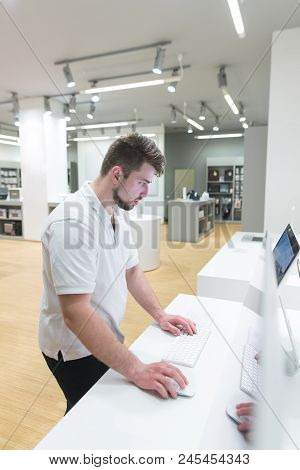 Man Uses A Computer In A Modern Electronics Store. Buyer Selects A Monoblock In A Light Technology S