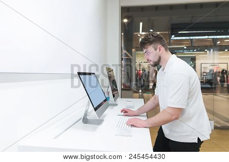 Man Uses A Computer In A Modern Electronics Store. Buyer Selects A Monoblock In A Light Technology S