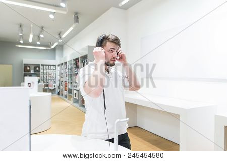 Handsome Man In A White T-shirt Tests Headphones In A Modern Tech Store. Buyers Listen To Music In H