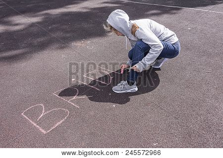 The Girl Writes Chalk On The Asphalt, In Windy Weather Is Dressed In A Jumper