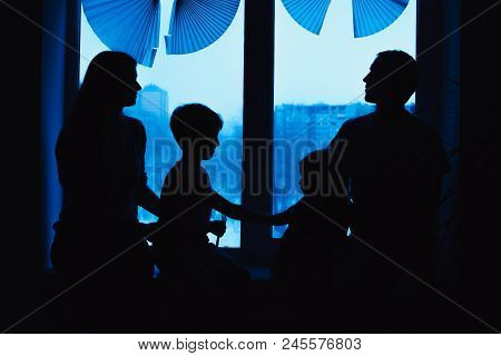 Parents And Kids Sitting Near Window And Having Fun At Home. Silhouette View. Happy Family On The Wi