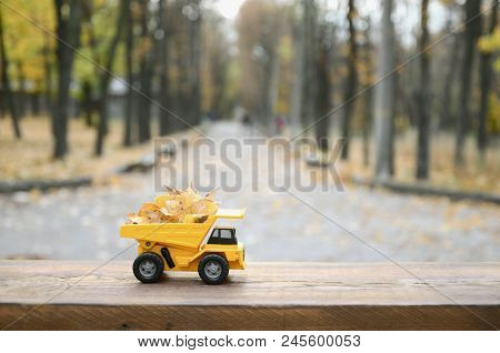 A Small Toy Yellow Truck Is Loaded With Yellow Fallen Leaves. The Car Stands On A Wooden Surface Aga