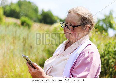 Elderly Woman In The Street Wearing Glasses Looks At The Mobile Phone.