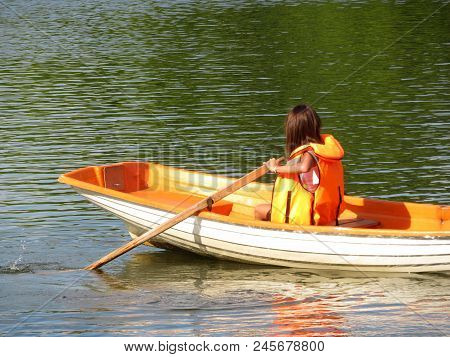 A Girl In A Life Jacket Floats On A Rowing Boat On A Summer Lake. Young Woman Rowing With Oars, Safe