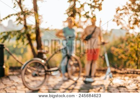 Blurred Background With Family Acticity Like Cycling And Scootering On The Hot Summer Evening.