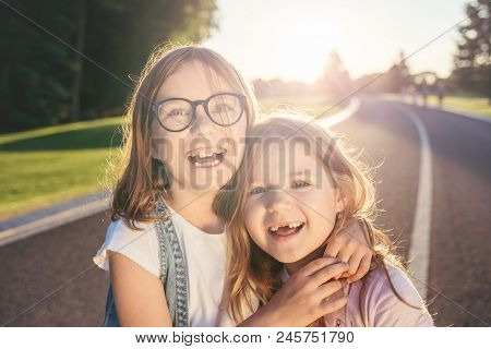 Two Joyfull Girls Standing On The Road, Hugging And Smiling Against The Sunset. Sincere Emotion. Gir