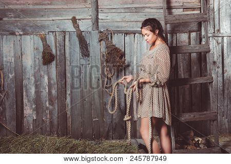 Young Girl In The Barn Holding Rope In Her Hands And Looking At It, Ladder And Brooms On Her Backgro
