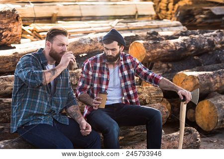 Two Bearded Loggers Sitting With Disposable Cups Of Coffee And Axe