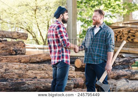 Side View Of Smiling Lumberjack With Tattooed Hand Holding Axe And Shaking Hands With Partner At Saw