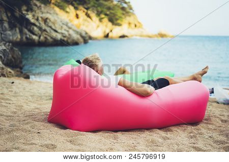 Young Hipster Relaxing On Coastline Beach On Inflatable Lazy Air Pouffe Sofa, Person Tourist Enjoy S