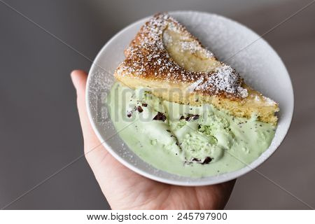 A Slice Of Apple Pie With Pistachio Ice Cream On A Wooden Background In A White Plate In A Woman's H