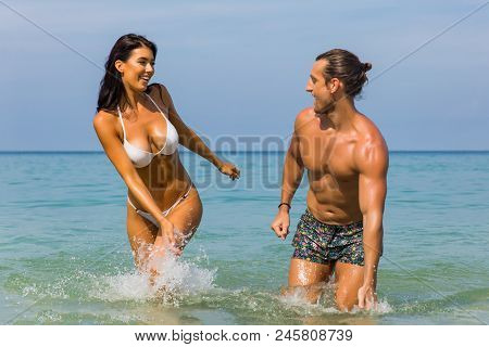 Happy Couple Laughing Together Holding Hands Running Having Fun Splashing Water In The Ocean Waves. 