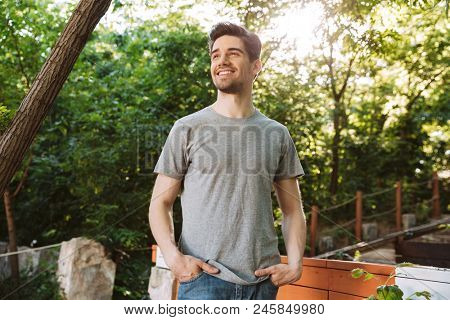 Pictire of Smiling brunette man in casual clothes looking away while holding arms in pockets outdoors