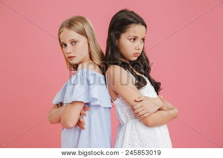 Image of two disappointed girls 8-10 years old wearing dresses standing back to back with arms crossed and expressing resentment isolated over pink background