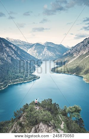 Happy female adventurer with backpack and arm raised enjoying amazing view to mountain lake in valley from top of mountain after summer hike - adventure, travel and hiking concept. Plansee, Austria