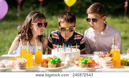 holidays, childhood and celebration concept - happy kids blowing out candles on birthday cake sitting at table at summer garden party