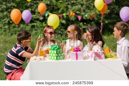 holidays, childhood and celebration concept - happy kids in sunglasses sitting at table on birthday party at summer garden and eating cupcakes