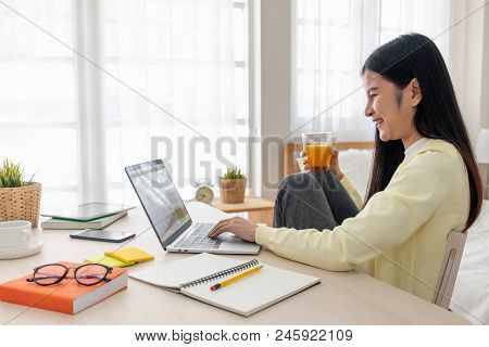 Asian Female Sit With Knees Up Use Social Media With Laptop On Table And Drink Orange Juice In Bedro