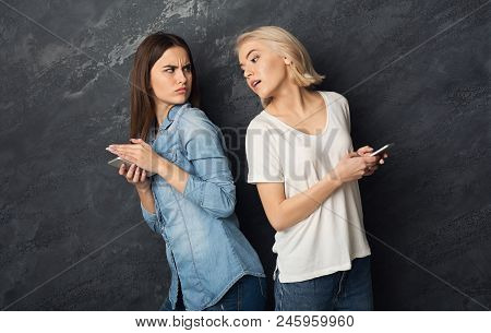 Girls Prying Into Each Other Phones At Gray Background. Spying Concept