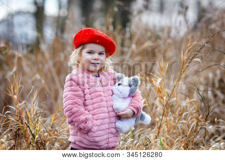 Outdoor Portrait Of Little Cute Toddler Girl In Pink Coat And Red Fashion Hat Barret Playing With So