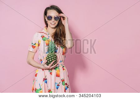 Beautiful Woman In Dress With Printed Friuts Posing With Pineapple On Pink Background. Pineapple In 