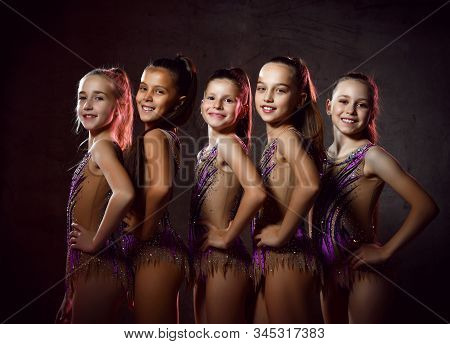 Team Of Young Smiling Girls Gymnasts In Purple Shiny Sport Body Posing Over Grey Background. Rhytmhi