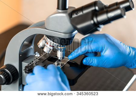 Male Laboratory Assistant Examining Biomaterial Samples In A Microscope. Cllose Up Hands In Blue Rub